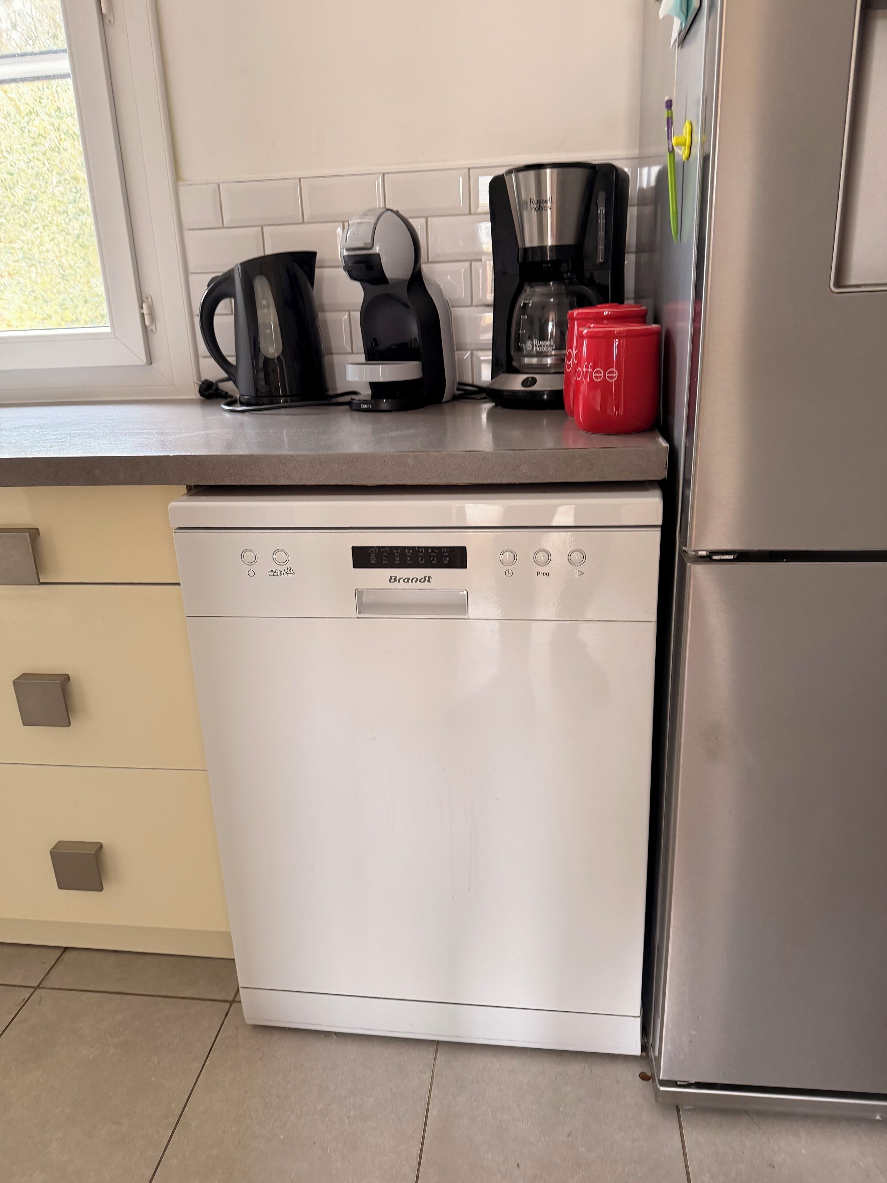 Una cocina con lavavajillas blanco, cafetera y una taza roja sobre la encimera.
