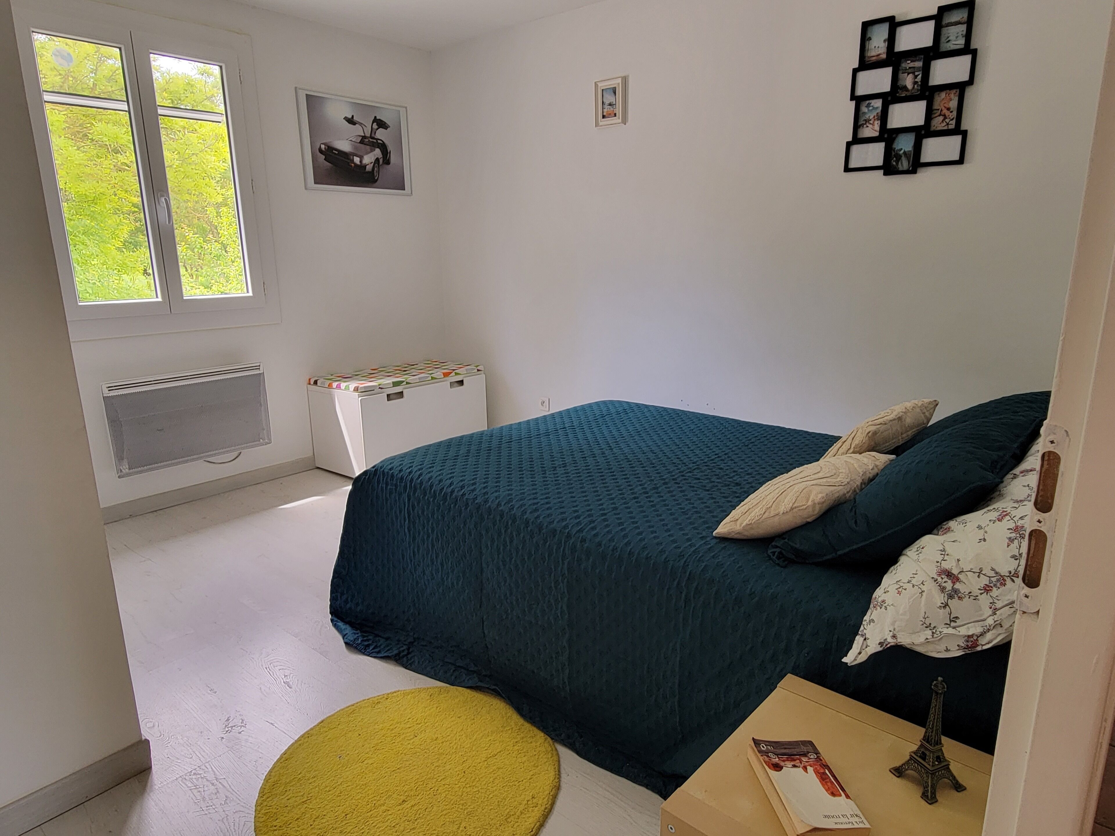 A bedroom with a bed, a window, a radiator, a small stool, and a wall with picture frames.