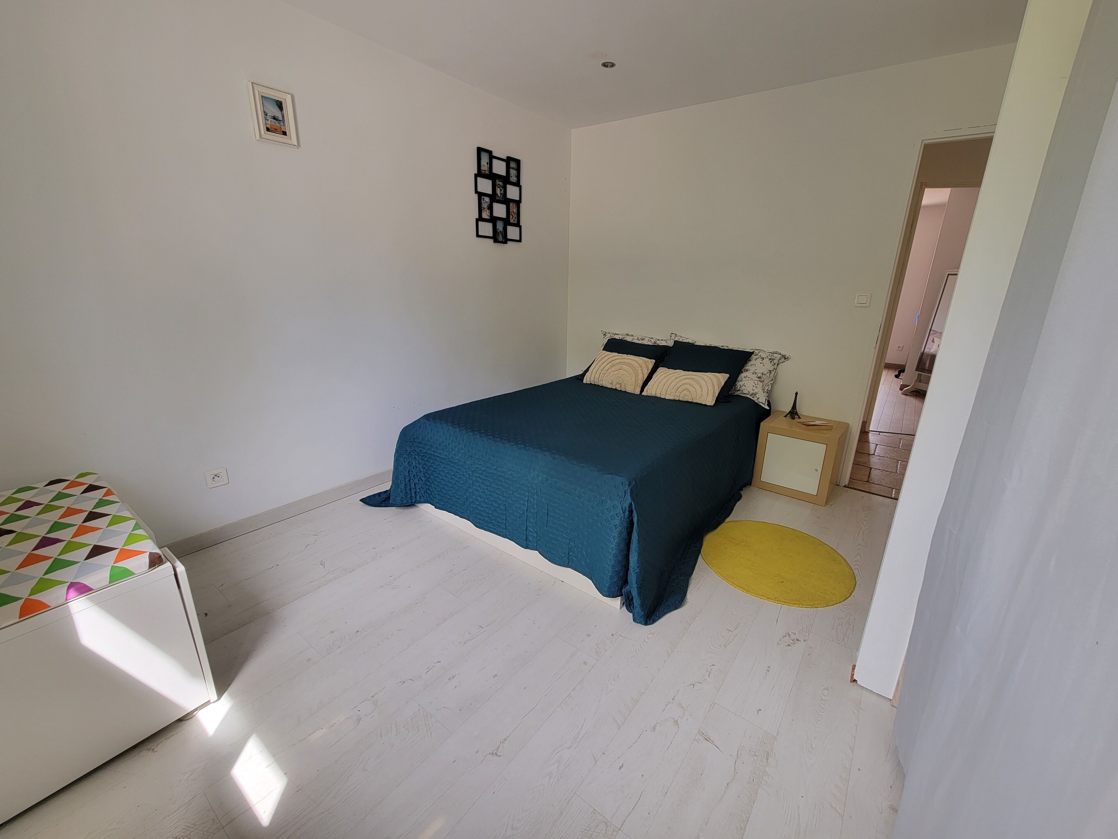A bedroom with a bed, a bedside table, a small stool, and a wall-mounted picture frame.