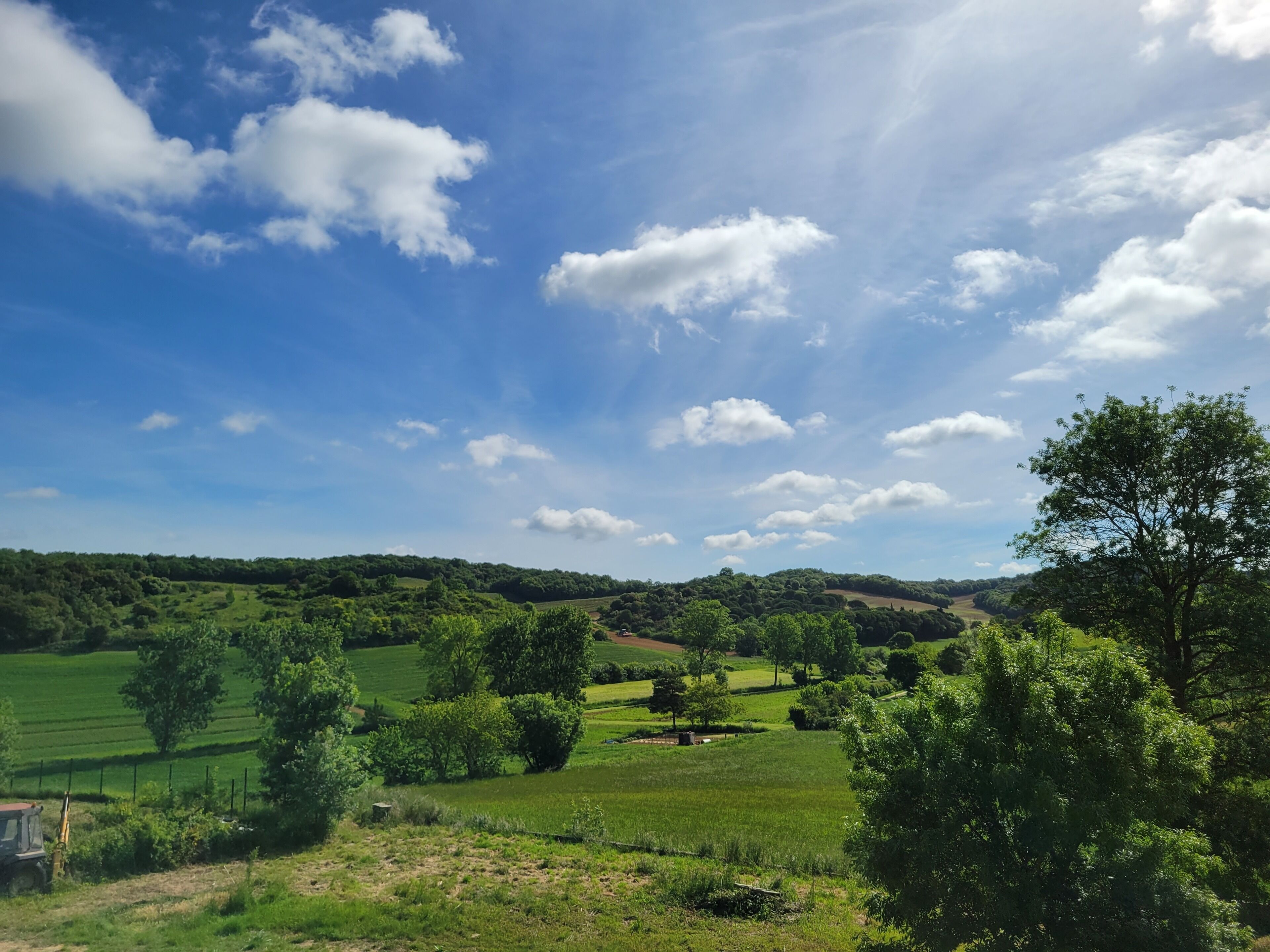 A rural landscape with rolling hills, green fields, and scattered trees under a clear blue sky with clouds.