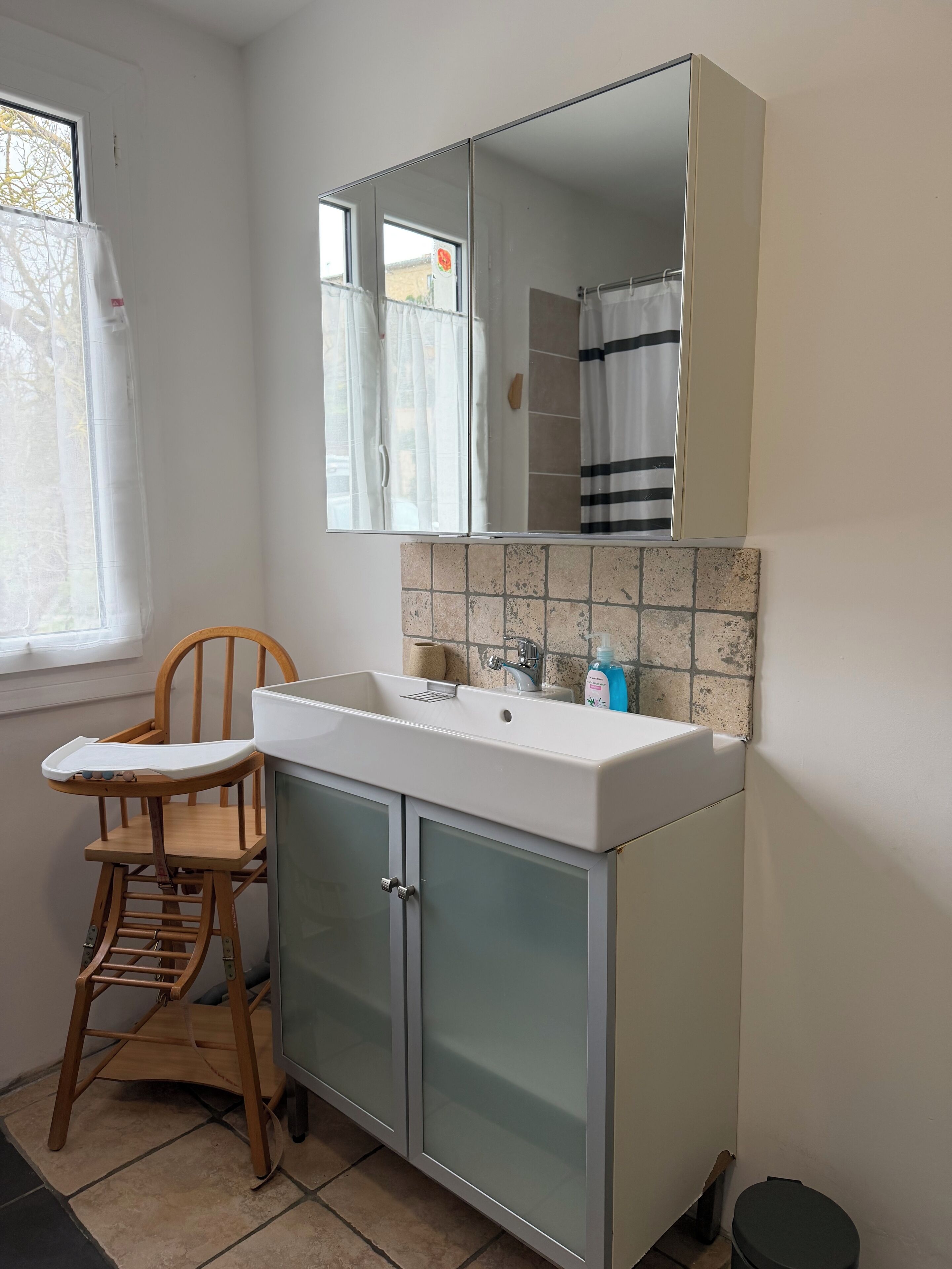 Een badkamer met een witte wastafel, een spiegel erboven, een houten stoel en een betegelde spatwand.