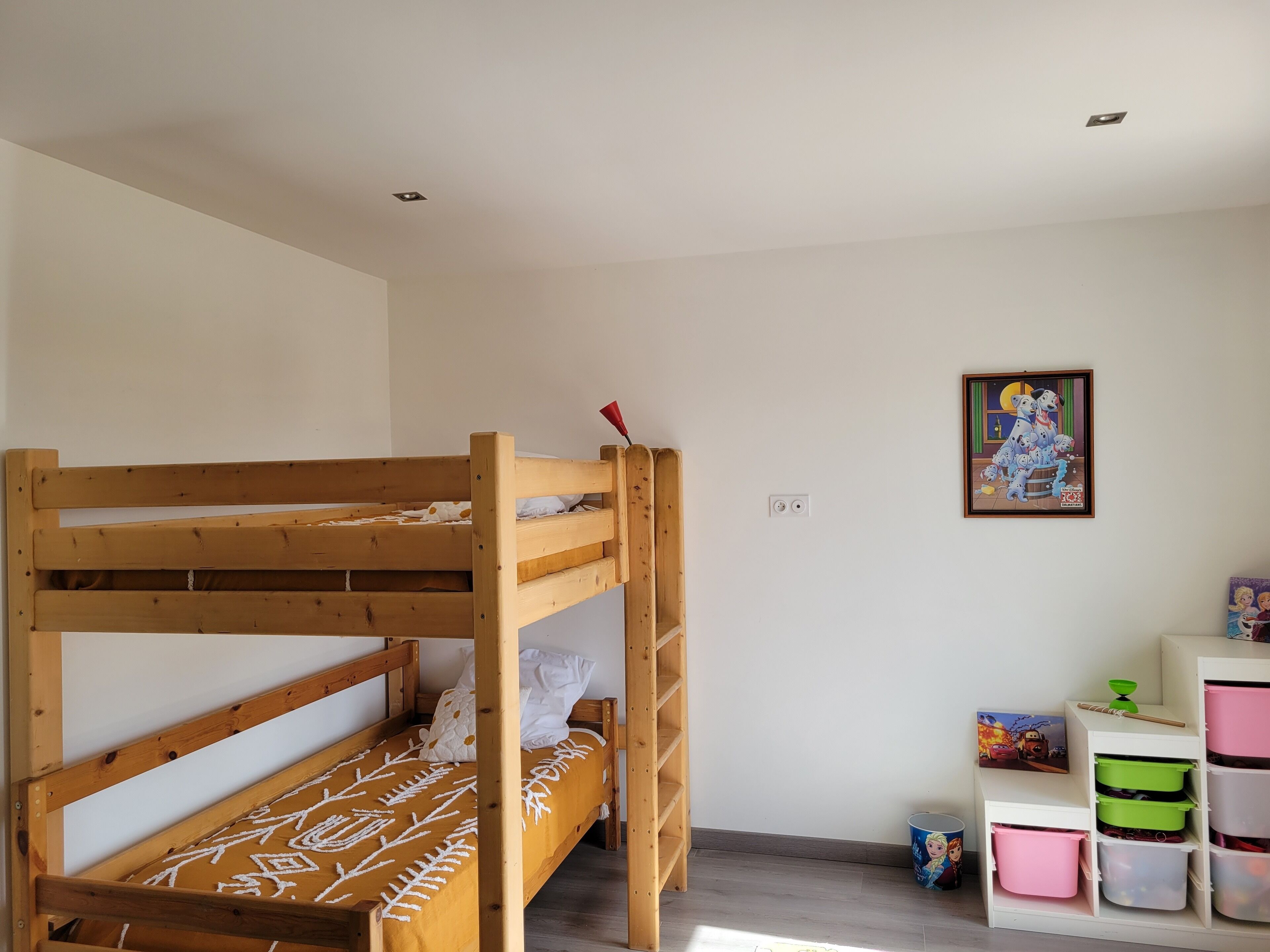Uma cama beliche com colcha estampada, uma mesa pequena com um objeto vermelho e unidades de armazenamento com caixas coloridas.