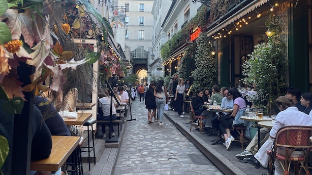 Une ruelle étroite pavée, bordée de terrasses et de cafés, où l’on peut voir des gens dîner et se promener.