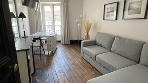 Un salon avec un canapé gris, un téléviseur fixé au mur, une table à manger avec des chaises et une fenêtre avec des rideaux.