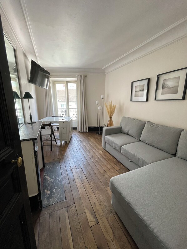Ein Wohnzimmer mit grauem Sofa, einem an der Wand befestigten Fernseher, einem Esstisch mit Stühlen und einem Fenster mit Vorhängen.