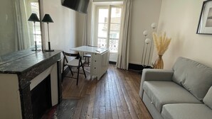 Un salon doté d’un plancher en bois, d’un canapé gris, d’un bureau avec une chaise, d’un téléviseur à écran plat fixé au mur et d’une fenêtre avec des rideaux.