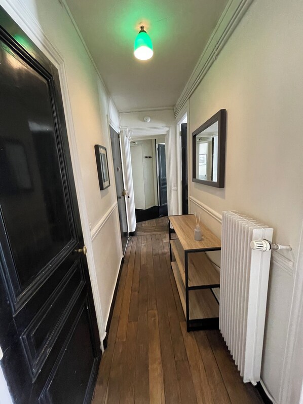 A hallway with wooden flooring, a radiator, a table with a bottle, and a mirror on the wall.