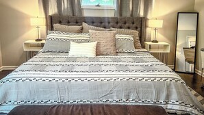 Ein ordentlich eingerichtetes Schlafzimmer mit einem großen Bett, zwei Nachttischen mit Lampen, einem Fenster mit Vorhängen und einem Spiegel an der Wand.