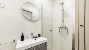 A modern bathroom with a white sink, a round mirror, a handheld showerhead, and a glass-enclosed shower. - Studio centre-ville Rennes proche Gare (Rennes)