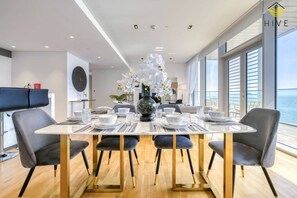 Un coin repas avec une table blanche dressée pour quatre personnes, entourée de chaises grises à revêtement en tissu et aux pieds dorés. Un grand vase rempli de fleurs blanches est placé au centre de la table.