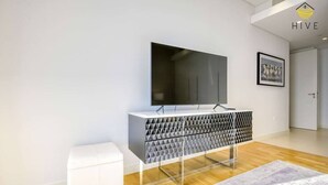 Een moderne woonkamer met een flatscreen televisie, een salontafel met een geometrisch patroon, een witte poef en een lijstfoto aan de muur.