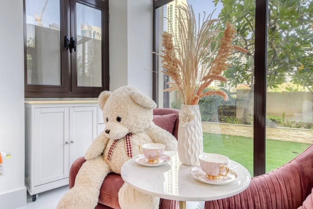 Een kamer met een grote witte knuffelbeer op een roze bank, een witte tafel met twee theekopjes, en een vaas met gedroogde planten.