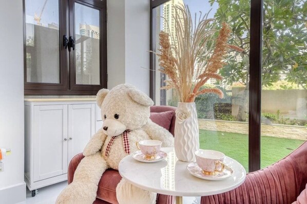 Une pièce avec un grand ours en peluche blanc sur un canapé rose, une table blanche avec deux tasses à thé et un vase contenant des plantes séchées.