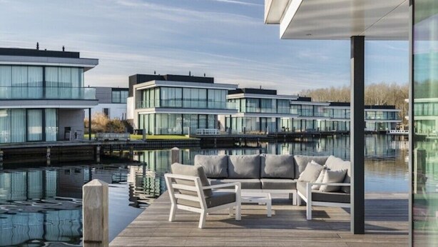 Ein modernes Gebäude mit Glasfassade, einer Holzterrasse mit Außenbestuhlung und einem ruhigen Wasserspiegel im Hintergrund.