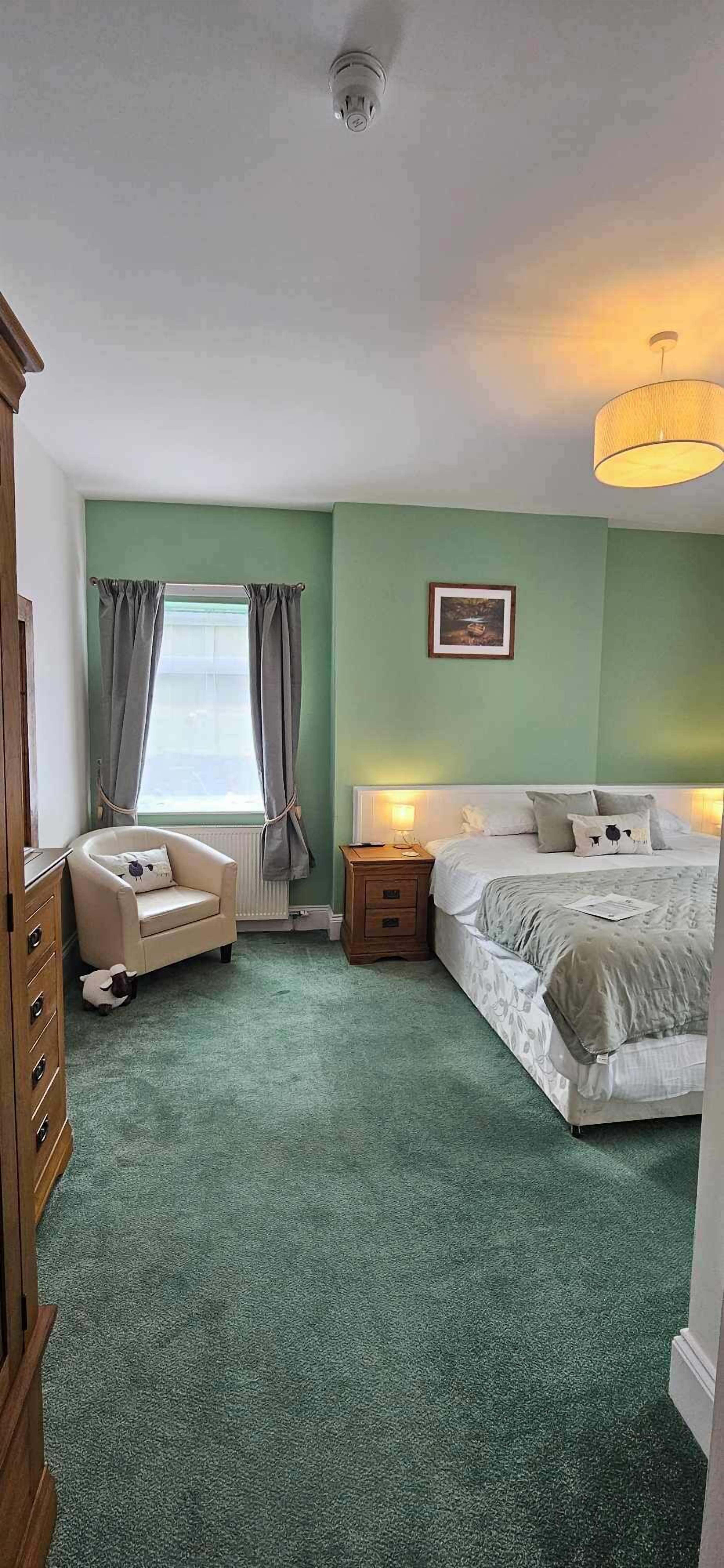 A bedroom with a bed, a chair, a dresser, a nightstand, a lamp, and a window with curtains.