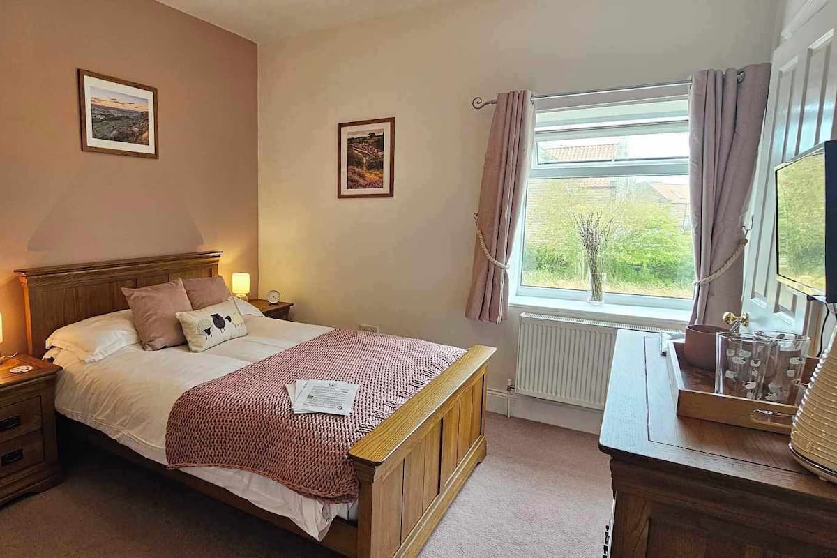 A bedroom with a wooden bed, bedside lamps, a window with curtains, and two framed pictures on the wall.