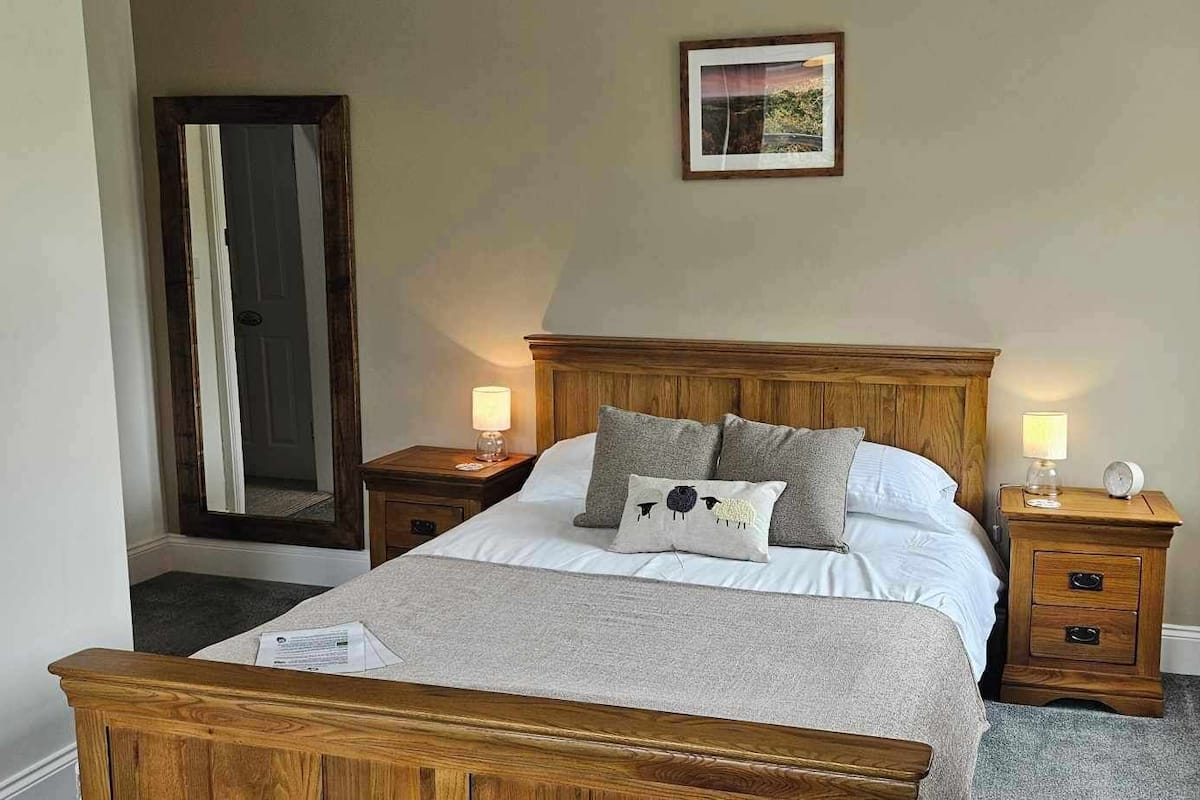 A neatly arranged bedroom with a wooden bed, two bedside tables, a nightstand, a mirror, and a framed picture on the wall.
