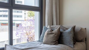 Een moderne slaapkamer met een groot raam, een bed met veel kussens en uitzicht op gebouwen en bomen.