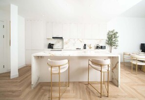A modern kitchen with white cabinetry, a marble countertop, two gold bar stools, and a small potted plant.