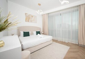 A modern hotel room with a large bed, a bedside table, a window with sheer curtains, and a wall-mounted artwork.