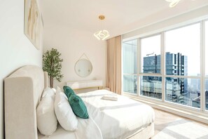 A modern bedroom with a large window, a bed with white bedding and a green pillow, a bedside table, a mirror, and a hanging light fixture.