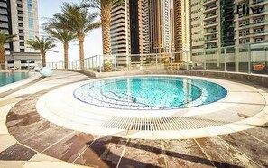 Uma piscina circular com uma fonte central, rodeada por um deck ladrilhado, e edifícios altos ao fundo.