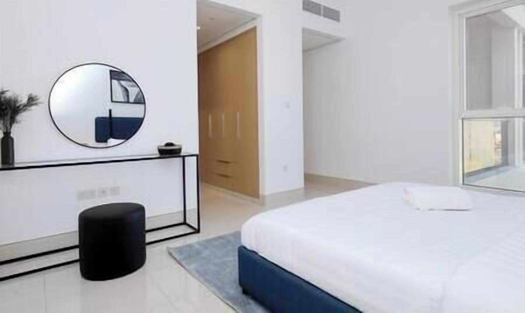 Une chambre à coucher moderne avec un lit, un miroir rond, un pouf noir et une armoire.