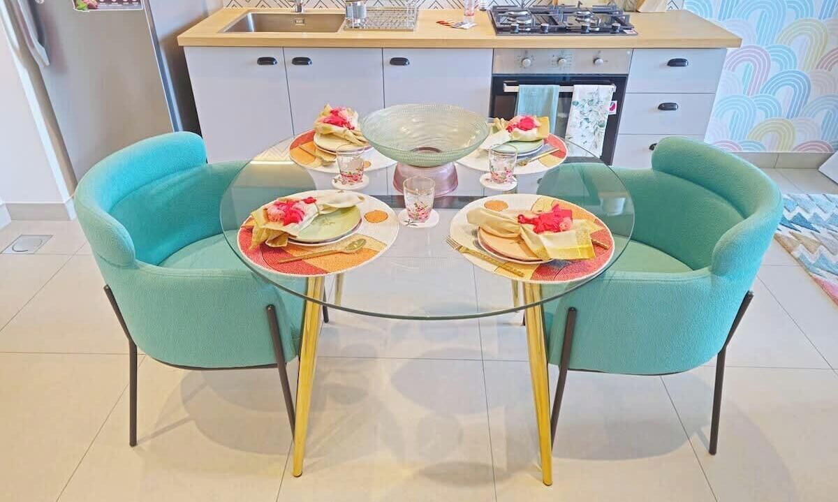 Un comedor con una mesa de vidrio rodeada de sillas color turquesa, un cuenco de vidrio en el centro y platos con servilletas dobladas.