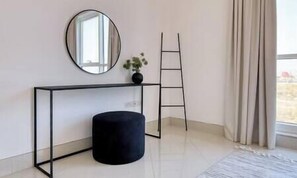 Ein Zimmer mit einer schwarzen Konsole, einem runden Spiegel, einem schwarzen Hocker, einer Leiter und einem Fenster mit Vorhängen.