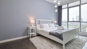 A bedroom with a large window, a white bed, and a fluffy rug. - Golf Course Views Apt W Pool & Fast Wifi (Dubai)
