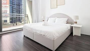 Une chambre moderne avec un grand lit, deux tables de chevet et une vue sur le paysage urbain, visible à travers une grande fenêtre.