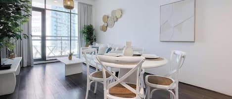 A modern dining area with a round table and white chairs, a large window with curtains, and a city view.
