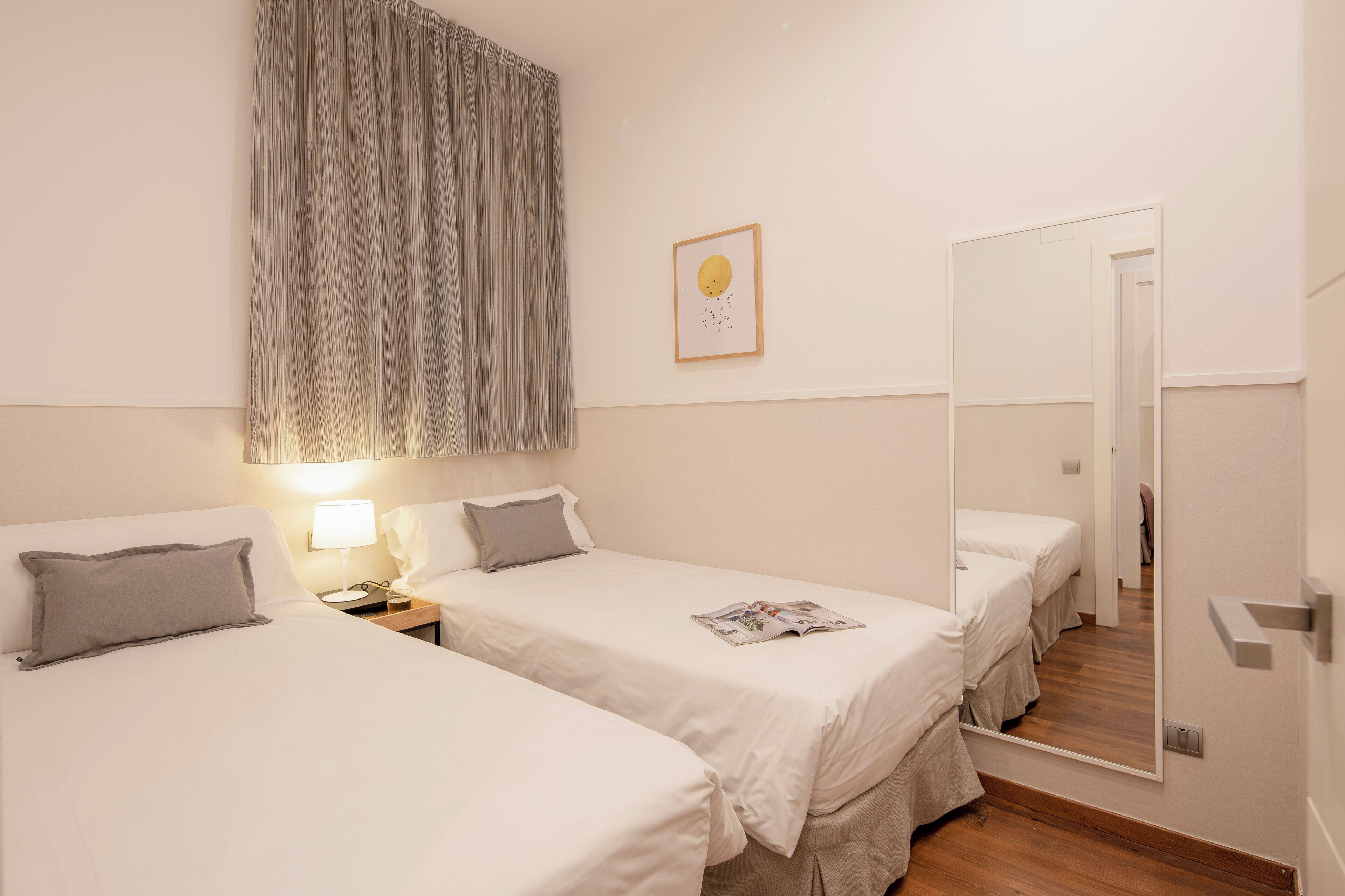 A hotel room with two single beds, a bedside table with a lamp, a large mirror, and a framed picture on the wall.