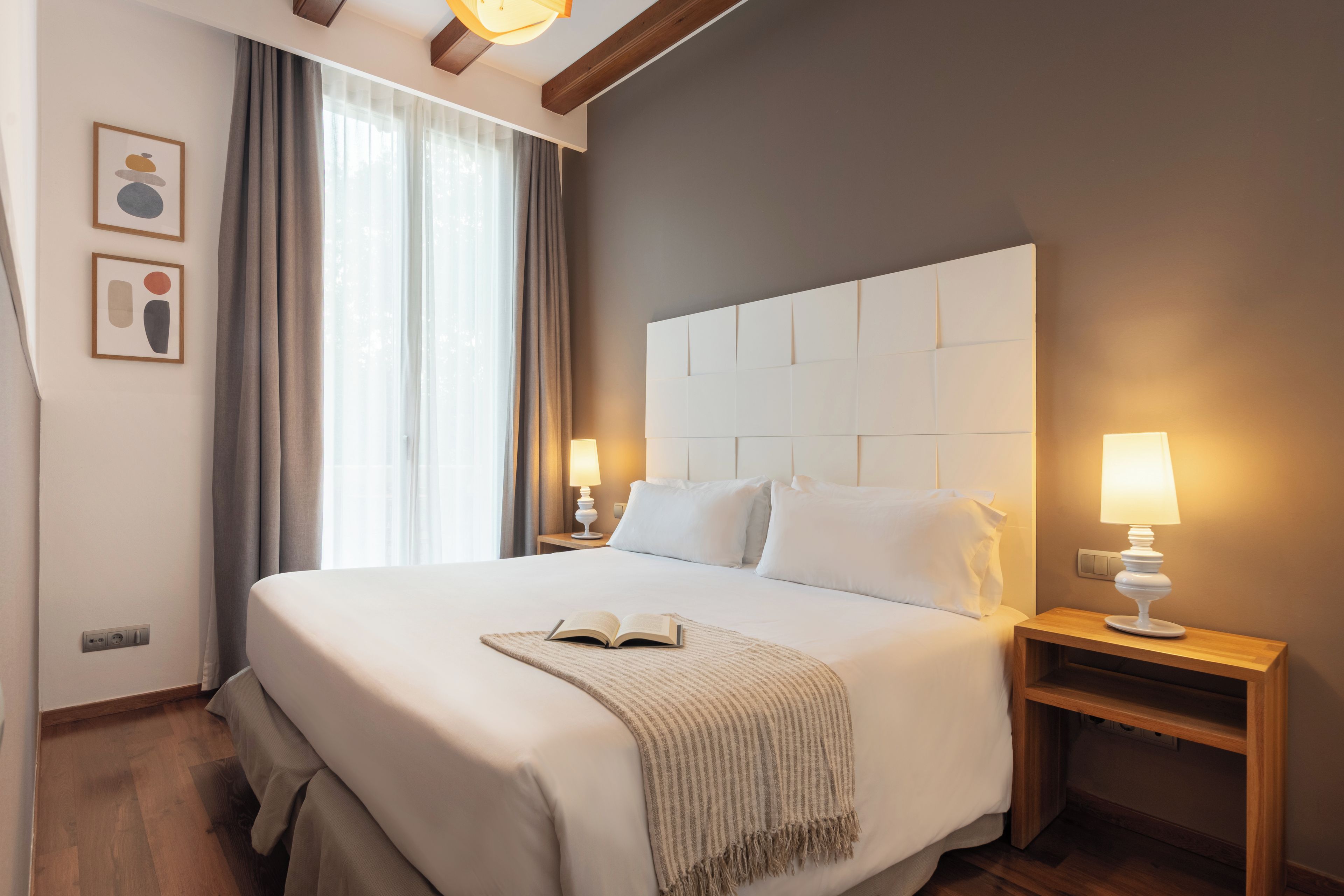 Quarto de hotel com uma cama grande, duas mesas de cabeceira com abajures, uma janela com cortinas e uma obra de arte na parede.