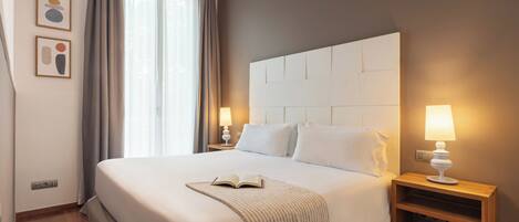 Quarto de hotel com uma cama grande, duas mesas de cabeceira com abajures, uma janela com cortinas e uma obra de arte na parede.