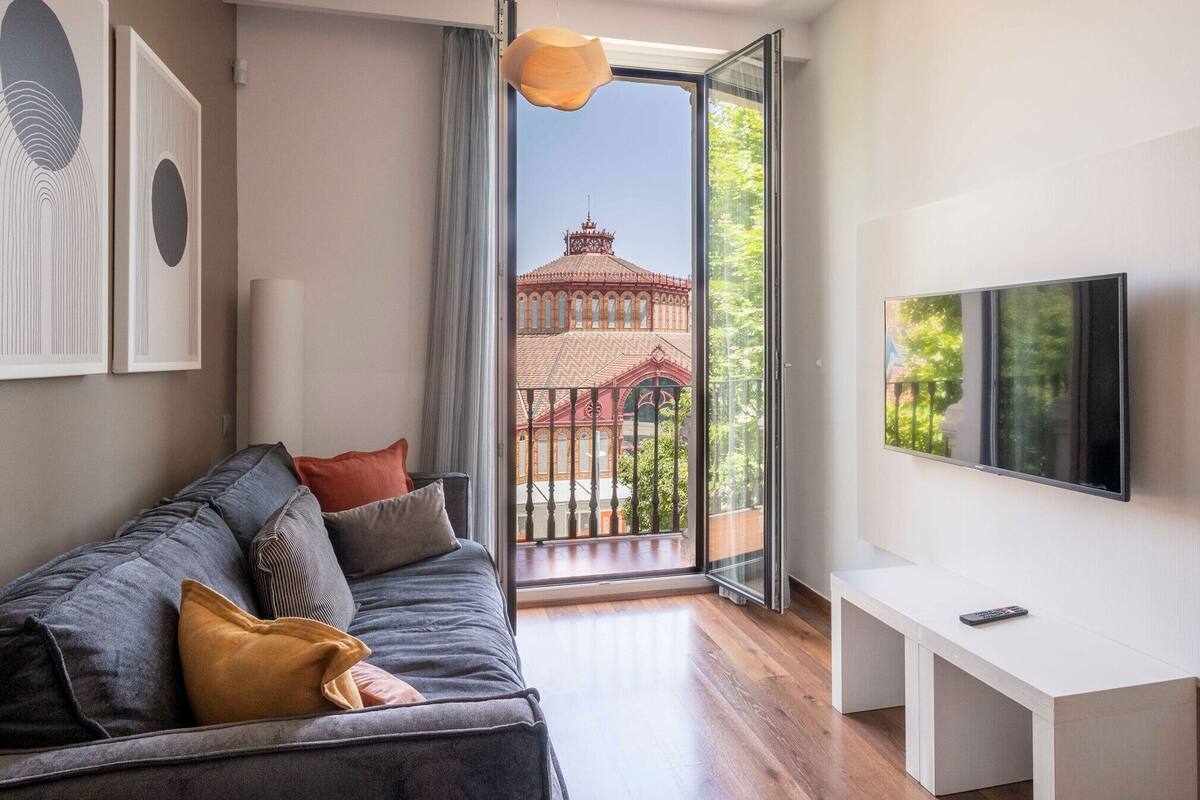 A modern living room with a sofa, a TV mounted on the wall, and a balcony with a view of a building.
