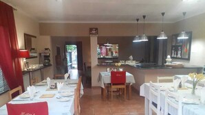 A dining area with multiple tables set for service, a kitchen in the background, and a red towel on a chair. - St Michelle Guest Lodge (Alberton)