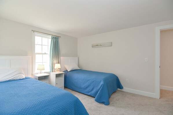 A bedroom with two beds, a window with curtains, a nightstand with a lamp, and a wall-mounted picture.