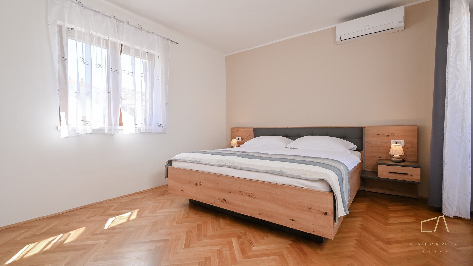 A bedroom with a wooden bed, bedside tables, a nightstand, and a window with sheer curtains.