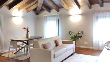 Un ampio soggiorno con soffitto in legno a vista, un divano bianco, un tavolo da pranzo con sedie e una grande finestra con tende leggere.