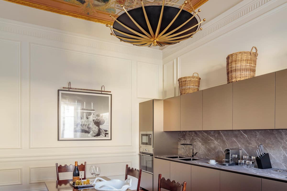 A dining area with a wooden table, chairs, and a chandelier. There is a kitchen area with cabinets and a microwave.