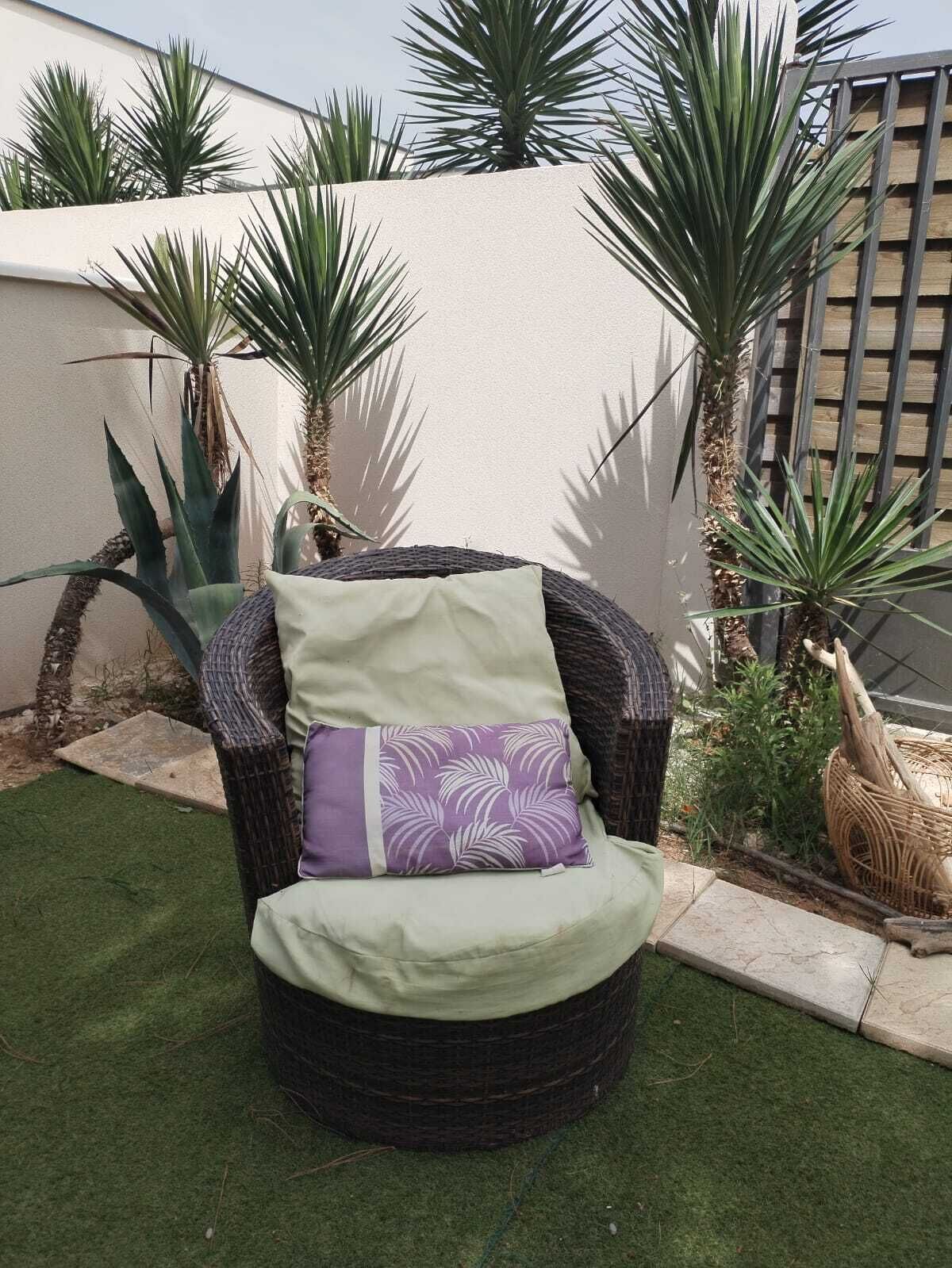 A wicker chair with a cushion and a decorative pillow in a garden area with artificial grass and palm plants.
