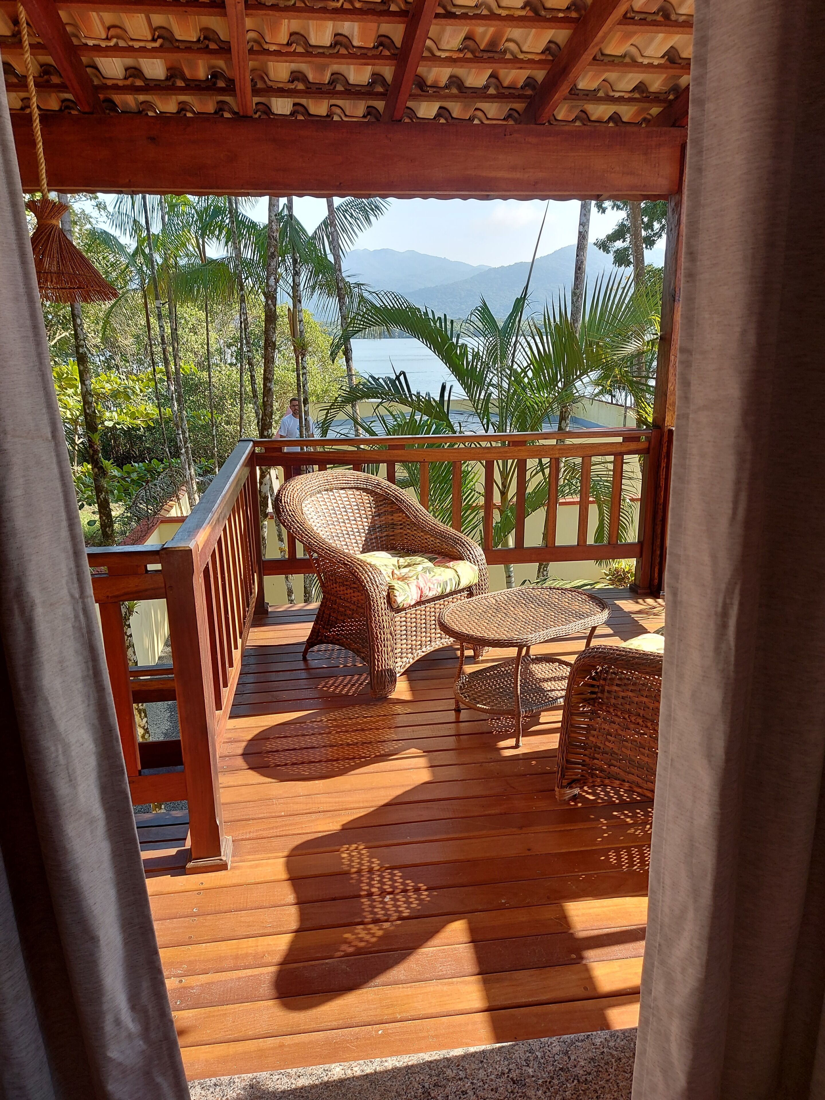 Um deck de madeira com mobiliário de vime, guarda-corpo e vista para o mar e montanhas.