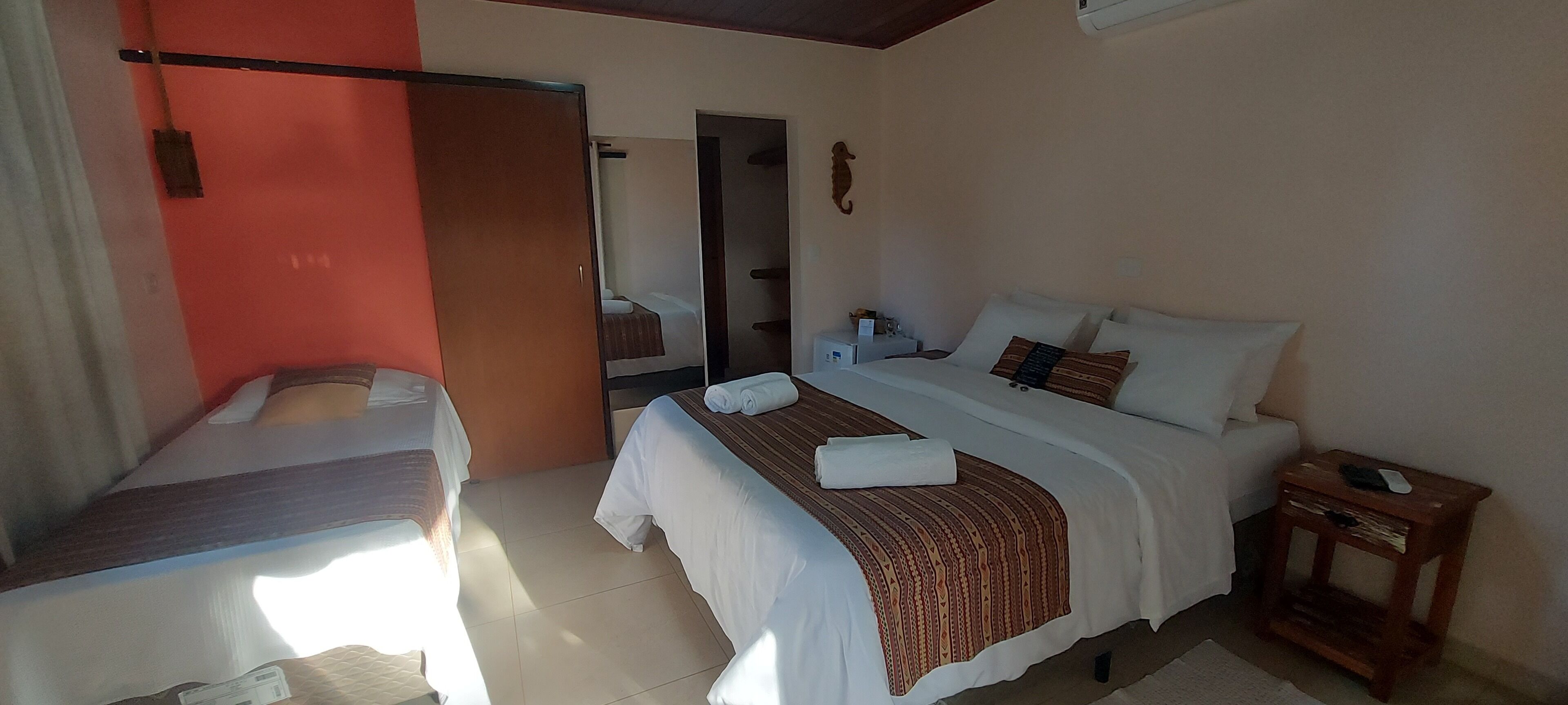 Quarto de hotel com duas camas, piso de madeira, espelho, pequena mesa e prateleira fixada na parede.
