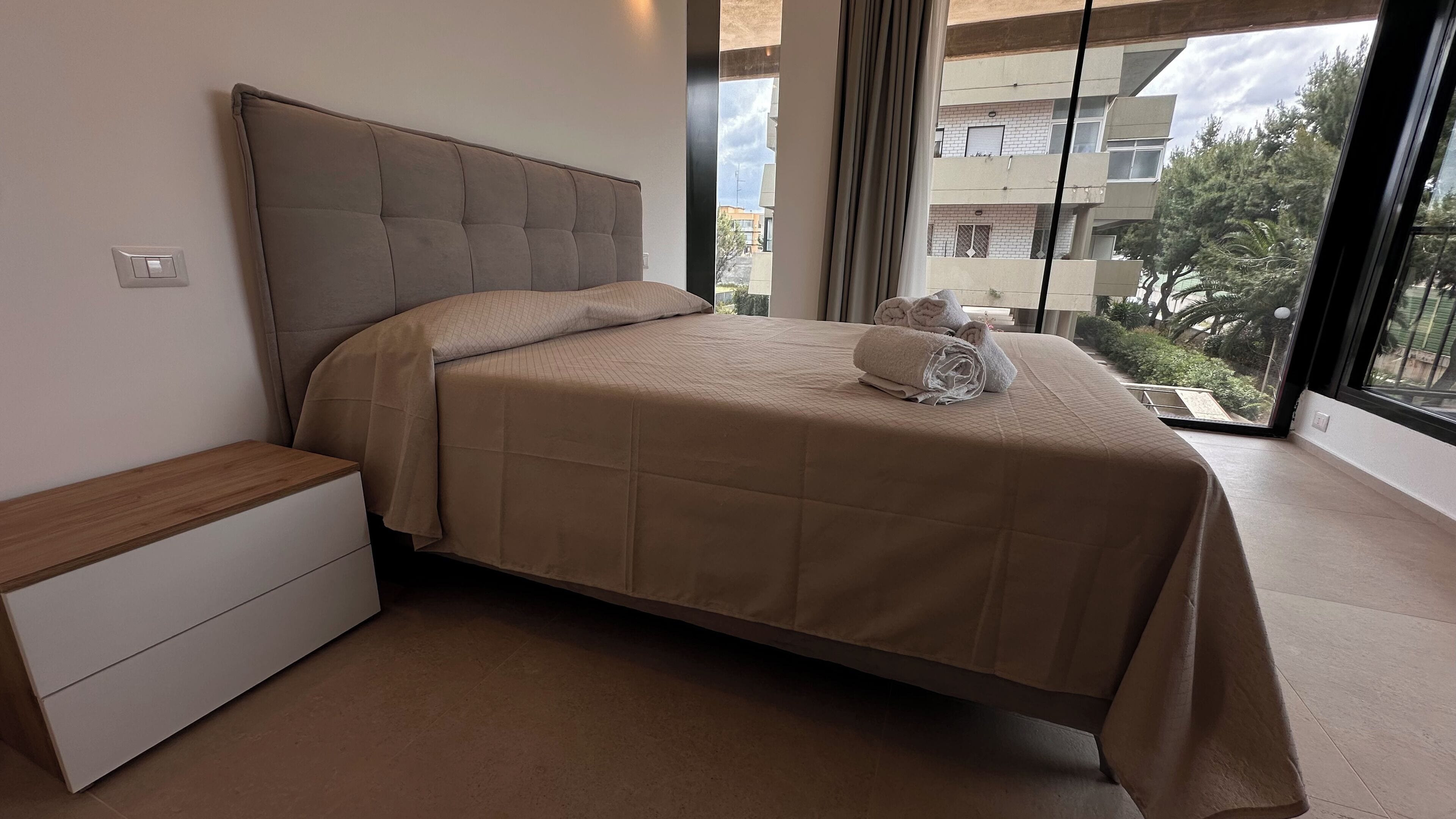 Une chambre avec un grand lit, une table de chevet et une vue sur les immeubles à travers de grandes fenêtres.