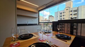 Ein Essbereich mit Tisch für zwei, eine Küche mit Spüle und Schränken sowie Blick auf Gebäude durch ein großes Fenster.