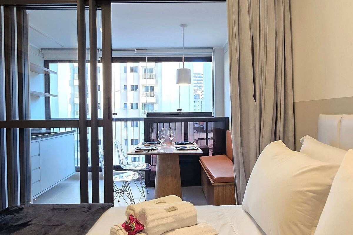 Habitación de hotel con cama, una pequeña mesa con sillas, balcón con vista a edificios y un amplio ventanal con cortinas.