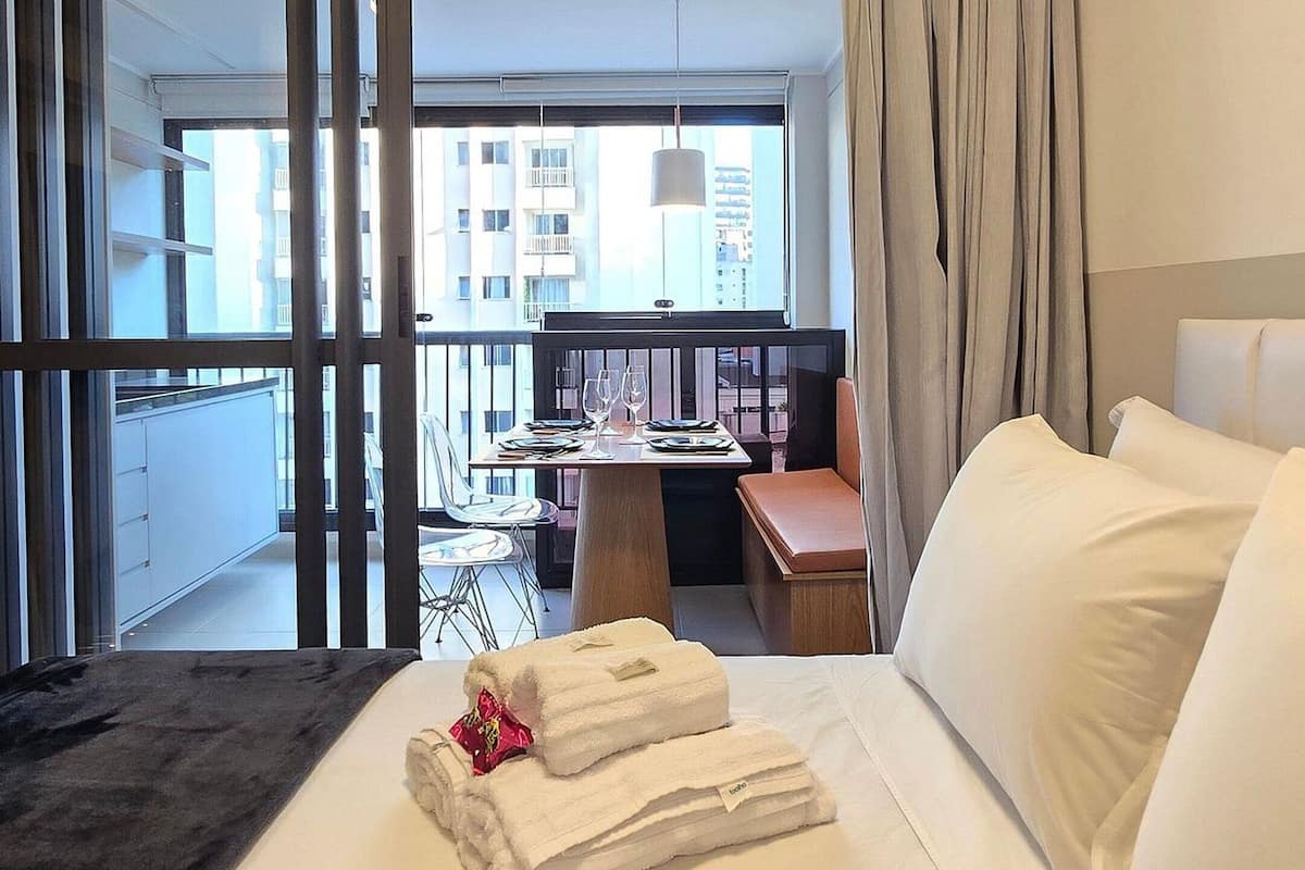 A hotel room with a bed, a small table with chairs, a balcony with a view of buildings, and a large window with curtains.