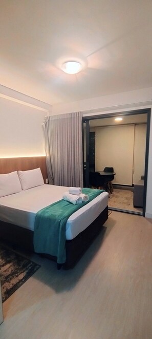 A hotel room with a bed, a green blanket, a curtain, a door leading to a dining area, and a ceiling light. - Charmosos Studios no coração de Sampa (São Paulo)