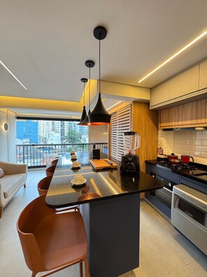 Eine moderne Küche mit einer zentralen Kochinsel, schwarzen Pendelleuchten und Blick auf die Stadt durch große Fenster.